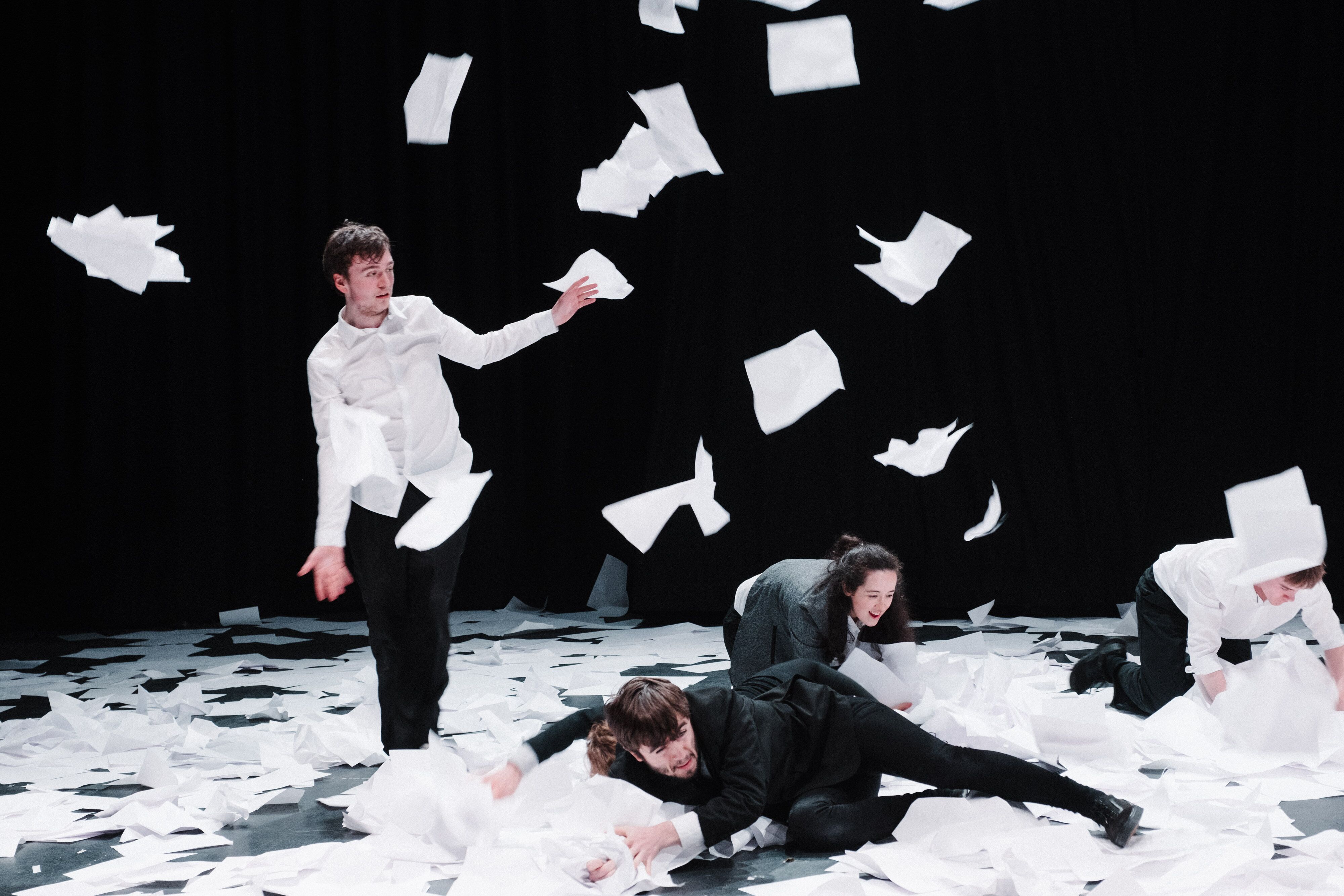 People on a stage in amongst a pile of white sheets of paper, throwing them around 